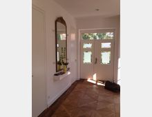 Heller Eingangsbereich mit einer weißen Tür, die vier teils mattierte Glasfenster enthält, und einem großen Wandspiegel mit verziertem Holzrahmen. Der Boden besteht aus Parkett, und auf einer schmalen Ablage unter dem Spiegel steht eine Vase mit Grünpflanzen.