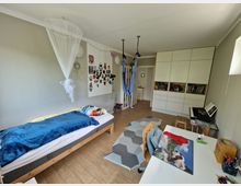 Schlicht eingerichtetes Kinderzimmer mit einem Einzelbett auf der linken Seite, einem Schreibtisch mit Stuhl rechts und einem großen weißen Einbauschrank im Hintergrund. An der Decke hängt ein weißes Moskitonetz, und im Raum befinden sich an der Wand bastelähnliche Dekorationen, eine blaue Hängematte sowie ein Keyboard auf einem kleinen Tisch.
