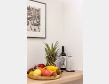 Eine minimalistische Anrichte mit einer goldenen Schale voller Obst (u. a. Ananas, Granatapfel, Bananen und Trauben) sowie einer Weinflasche und zwei Weingläsern. An der weißen Wand hängt ein gerahmtes Schwarz-Weiß-Bild einer historischen Stadtszene.