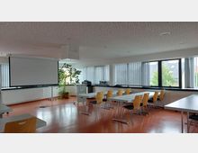 Ein moderner Seminarraum mit rechteckigen Tischen und Holzstühlen, einer heruntergezogenen Projektionsleinwand, großen Fensterfronten mit vertikalen Jalousien und einer Topfpflanze in der Ecke. Der Boden ist rot, und an den Wänden befinden sich Heizkörper.