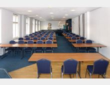 Ein Tagungsraum mit mehreren Reihen von rechteckigen Holztischen und gepolsterten blauen Stühlen. Der Raum verfügt über große Fenster an der linken Wand und hat einen blauen Teppichboden sowie eine helle Decke mit eingebauten Deckenleuchten.