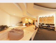 Ein modernes Büro mit einer offenen Raumgestaltung, bestehend aus einer Holzplattform mit Sitzkissen und Sitzsäcken auf der linken Seite und einer Arbeitszone mit Schreibtischen und Computerbildschirmen rechts. Die Backsteinwand im Hintergrund und die dekorativen, orangefarbenen Lampen betonen das industrielle Design.