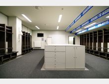 Umkleideraum mit dunklen Holzbänken und offenen Schließfächern an beiden Seitenwänden. In der Mitte steht eine weiße Kommode, darüber weiße Decken mit integrierter Beleuchtung; blaue Banner mit Motivationssprüchen zieren die Decke.