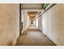 Langer, schmaler Korridor mit weißen Wänden, einer sichtbaren Holzdecke und freiliegenden Rohren an der Decke und den Seitenwänden. Der Boden ist aus Beton, und am Ende des Korridors ist eine geschlossene Tür sichtbar.