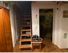 Innenraum eines Wohnbereichs mit Holzboden und sichtbaren Holzbalken. Im Vordergrund eine schmale Holztreppe, rechts ein Durchgang zu einem weiteren Raum mit Schreibtisch und Fenster, links ein Schrank und mehrere helle Wände.