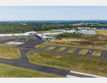 Luftaufnahme eines kleinen Flughafens in ländlicher Umgebung, mit mehreren Hangars, einer Luftverkehrskontrollstelle und geparkten Flugzeugen auf dem Vorfeld. Im Hintergrund sind Wälder, Wiesen sowie einige industrielle und landwirtschaftliche Gebäude zu sehen.
