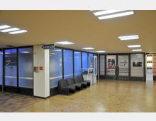 Ein Innenbereich eines Bürogebäudes mit hell erleuchtetem Flur, Glastüren und einer durchgehenden Fensterfront mit blau getöntem Glas. Im Vordergrund stehen schwarze Sitzstühle und ein kleines Ausstellungsregal, während an der rechten Wand Klingelschilder und ein Hinweis auf Räume L223–L229 sichtbar sind.