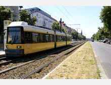 Gelbe Straßenbahn der Linie M2 an einer Haltestelle in einer urbanen Umgebung mit Wohnhäusern im Hintergrund, daneben verlaufen Fahrspuren und parkende Autos; die Gleise sind von Rasenstreifen und Bäumen gesäumt.