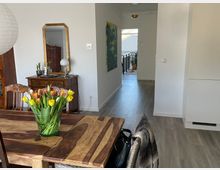 Wohnzimmer mit hellem Holzfußboden, einem Esstisch aus dunklem Holz, auf dem ein Glas mit bunten Tulpen steht. Im Hintergrund ein antiker Holzschrank, ein goldgerahmter Spiegel und ein Durchgang zu einem Balkon mit Blick auf ein Dach.