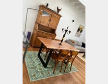 Essbereich mit Holztisch und vier passenden Holzstühlen auf einem gemusterten, grünen Teppich. Im Hintergrund steht eine große, antik wirkende Holzkommode, daneben eine Stehlampe, und an der Wand hängt ein abstraktes Gemälde.