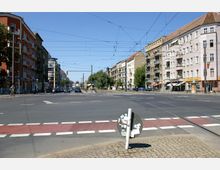 Straßenkreuzung in einer Stadt mit mehrstöckigen Wohn- und Geschäftsgebäuden an beiden Seiten. Oberleitungen für Straßenbahnen sind sichtbar, ebenso wie Fahrbahnen, Gehwege, Fußgängerüberwege und parkende Autos.