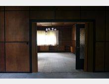 Ein leerer Raum mit Holzvertäfelung an Wänden und Decke, sichtbarer Übergang durch eine Türöffnung. Im Raum befindet sich ein Sideboard vor einem Fenster mit halbtransparenten Vorhängen, durch das gedämpftes Tageslicht einfällt.