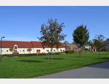 Große Wiese mit einzeln stehenden Bäumen im Vordergrund, dahinter ein längliches Gebäude mit heller Fassade und rotem Ziegeldach. Im Hintergrund weitere Gebäude und eine Gartenanlage mit Wegen und Bepflanzung unter klarem, blauem Himmel.