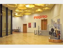 Eingangsbereich eines Veranstaltungs- oder Kinogebäudes mit Holzfußboden, Wänden aus hellem Mauerwerk und gelben, wabenförmigen Deckenleuchten. Im Raum befinden sich Türen aus Holz, Schaukästen mit alten Filmprojektoren und Plakaten sowie der Schriftzug „Fezino“ an der Wand.