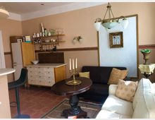 Wohnbereich mit klassisch-eleganter Einrichtung: Eine kleine Küche mit offenen Holzregalen und einem weißen Schrank auf der linken Seite, daneben ein Sitzbereich mit einem Ledersofa, einem Polstersofa, dekorativen Kissen, einem runden Holztisch und einem Kronleuchter. Wände in Beige mit goldgerahmtem Bild und Leuchten.