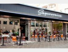 Außenansicht eines Cafés oder Restaurants mit dem Namen „Supersonico“, erkennbar durch ein beleuchtetes Schild. Vor der Fassade mit großen Fenstern stehen mehrere Tische und Holzstühle auf einem Gehweg, im Innenbereich sind Gäste und Personal sichtbar.