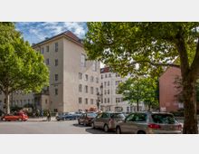 Straßenszene in einer städtischen Umgebung mit einem beigen Gebäude, das mit „Puppentheater Berlin“ beschriftet ist. Im Vordergrund stehen mehrere geparkte Autos, während Bäume und ein kleiner Platz für eine grüne Umgebung sorgen.