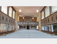 Innenraum einer historischen Kirche mit hölzernen, hellgrau gestrichenen Sitzbänken und umlaufenden Emporen, die mit dekorativen Wappendarstellungen verziert sind. Im Hintergrund befindet sich die Orgelempore mit einer Inschrift darüber; die Raumgestaltung ist schlicht mit hellen Wänden und Hängeleuchten.