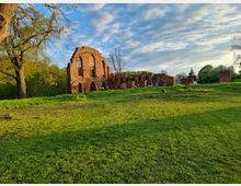 Ruine eines historischen Backsteingebäudes inmitten einer grünen Wiese, umgeben von Bäumen und unter einem teils bewölkten Himmel bei tiefstehender Sonne. Die Überreste zeigen Spitzbögen und bestehende Mauern, die auf eine frühere kirchliche oder gotische Architektur hindeuten.