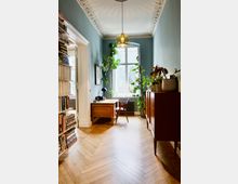 Ein heller Raum mit hölzernem Fischgrätenparkett, einem hohen Fenster und einer mit Pflanzen dekorierten Einrichtung. Links befinden sich Bücherregale, rechts ein Sideboard aus Holz, und am Fenster steht ein schlichter Schreibtisch mit Stuhl.