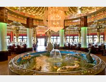 Innenraum eines imposanten Restaurants im asiatischen Stil mit dekorativen Säulen, vergoldeten Ornamenten und einem auffälligen Kronleuchter. Im Vordergrund befindet sich ein runder, mit Figuren und Fischen geschmückter Brunnen, umgeben von gedeckten Tischen.