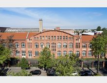 Backsteinfassade eines historischen Industriebaues mit großen, weißen Sprossenfenstern und einem roten Satteldach. Im Vordergrund stehen Bäume und geparkte Autos an einer Straße, dahinter ist der Ziegelbau mit einem Schornstein und einer urbanen Umgebung im Hintergrund zu sehen.