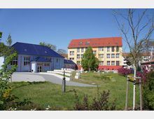 Zwei Gebäude in einem parkähnlichen Umfeld, ein kleineres blaues Gebäude mit dunklem Dach im Vordergrund und ein größeres gelb-rotes Gebäude mit rotem Ziegeldach im Hintergrund. Dazwischen befinden sich grüne Rasenflächen, Bäume, Büsche und ein gepflasterter Weg.