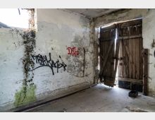 Ein verlassener Raum mit bröckelnden, teils mit Graffiti bedeckten Wänden; eine hölzerne, beschädigte Doppeltür lässt Licht von außen herein. An der linken Wand wächst Efeu neben einem kleinen Fenster.