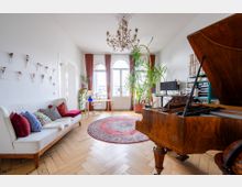 Helles Wohnzimmer mit Holzboden, einem weißen Sofa mit bunten Zierkissen, einem geöffneten Holzflügel, großen Fenstern mit roten Vorhängen und mehreren Pflanzen. Eine runde Teppichfläche und ein Kronleuchter ergänzen das klassische Ambiente.