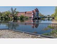 Ansicht eines Flusses mit einer Brücke im Hintergrund und einem historischen roten Backsteingebäude mit Satteldach dahinter. Im Vordergrund befindet sich ein Geländer, das eine gepflasterte Uferpromenade begrenzt, umgeben von Bäumen und Schilf.