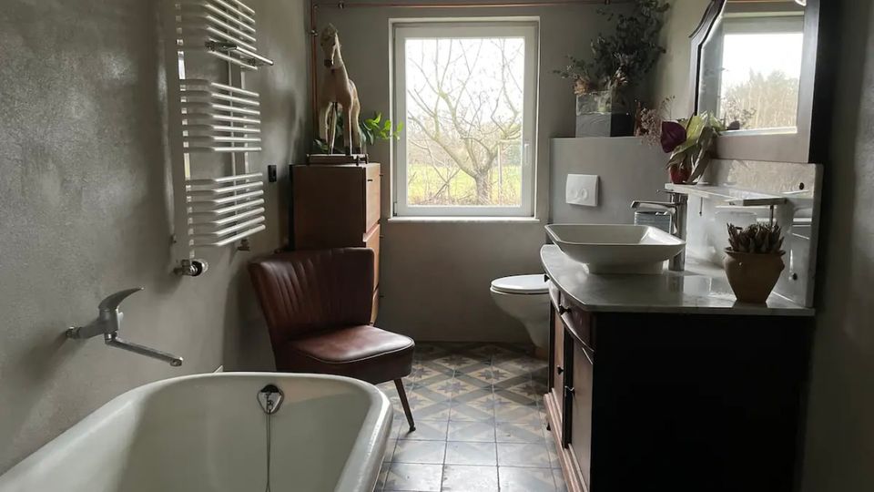 Helles Badezimmer mit freistehender Badewanne, einem Waschtisch mit Aufsatzwaschbecken und Spiegel, sowie einem Fenster, das auf eine grüne Landschaft blickt. Der Raum verfügt über graue Wände, gemusterte Bodenfliesen und dekorative Elemente wie Pflanzen und eine Vintage-Holzkommode.