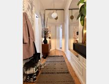 Schmaler Flur in einer Wohnung mit Holzdielenboden und hellem Wandanstrich, verziert mit floralen Tapetenmustern. Links befindet sich eine Garderobe mit Jacken und einem Schuhregal, rechts ein großer Spiegel; im Hintergrund stehen ein kleines Holzmöbelstück mit dekorativer Vase und mehrere weiße Türen.