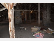 Dachboden eines alten Gebäudes mit sichtbaren Holzträgern, massiven Steinsäulen und teils beschädigtem Boden. Im Raum liegen verstreut Kartons, eine Kunststoffkiste, und andere Gegenstände.