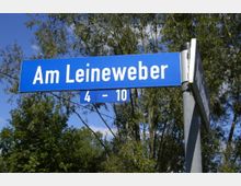 Blaues Straßenschild mit der Aufschrift „Am Leineweber 4-10“ vor einem Hintergrund aus Bäumen und blauem Himmel.