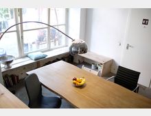 Heller Büroraum mit einem großen Holztisch, darauf eine Schale mit Obst, umgeben von modernen Stühlen. Im Hintergrund sind ein großes Fenster mit Blick nach draußen, eine silberne Stehlampe sowie eine schlichte weiße Tür mit Toilettenbeschilderung sichtbar.