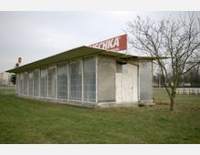 Ein rechteckiges, einfaches Gebäude mit durchsichtigen Kunststoffwänden und einem flachen Metalldach, auf einem grasbewachsenen Platz. An der Oberseite des Gebäudes befindet sich ein Werbeschild mit rotem Hintergrund und weißer Schrift.