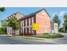 Straßenszene mit einem roten Ziegel- und einem hellrosa-verputzten Wohngebäude an einer Kreuzung. Im Vordergrund steht ein gelbes Verkehrsschild mit Wegweisern zu umliegenden Orten, umgeben von Gehweg, Fahrbahn und einigen Bäumen.