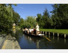 Personen sitzen in einem flachen Holzboot, das von einem Steuermann mit einer langen Stange durch einen schmalen Kanal gelenkt wird, umgeben von grünen Bäumen, Rasenflächen und einem kleinen Holzgebäude im Hintergrund.