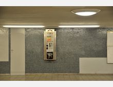 Ein Bild eines U-Bahnhofs oder Untergrundbereichs mit blauer Wand aus kleinen Mosaikfliesen und einem Wandtelefon in der Mitte. Der Boden besteht aus beige-grauen Fliesen, und Deckenleuchten sorgen für indirekte Beleuchtung.