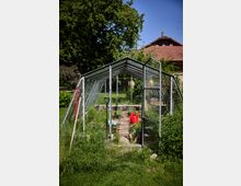 Kleines Gewächshaus mit Aluminiumrahmen und transparenten Glas- oder Kunststoffplatten in einem Garten, mit geöffnetem Eingang und schmalem Mittelweg. Im Inneren stehen Pflanztöpfe und eine rote Gießkanne; im Hintergrund sind Rasen, große Bäume und ein Haus mit rotem Ziegeldach zu sehen.
