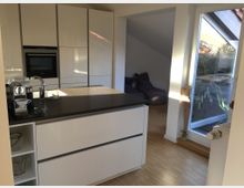 Moderne Küche mit weiß lackierten Schränken, einer dunklen Arbeitsplatte und integriertem Backofen. Im Hintergrund ein weiterer Raum mit Holzfußboden, Sitzsäcken und einer großen Fensterfront, die viel Tageslicht hereinlässt.