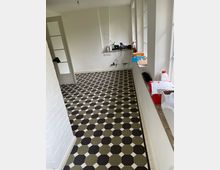 Zimmer mit hellem Anstrich und einem Boden aus schwarz-weiß-beige gemusterten Fliesen in achteckigem Design. Auf der rechten Seite befinden sich große Fenster und eine Ablage mit Gegenständen, während links eine halb geöffnete Tür zu sehen ist.