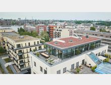 Wohngebiet mit modernen Mehrfamilienhäusern und begrünten Flachdächern, darunter ein Gebäude mit einer Dachterrasse, Sitzgelegenheiten und Glasfassaden. Im Hintergrund sind weitere Wohngebäude, Grünflächen und ein Fluss zu sehen.