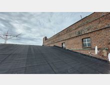 Flachdach aus schwarzem Material vor einer ziegelroten Backsteinwand mit Fenstern, Kabeln und einer Satellitenschüssel; im Hintergrund ein grauer, bewölkter Himmel und ein Teil einer Metallantenne.