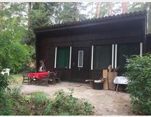 Ein simples, dunkles Ferienhaus aus Holz mit flachem Wellblechdach, umgeben von dichtem Wald. Vor dem Gebäude stehen ein Tisch mit roter Tischdecke und Holzstühlen sowie gestapelte Holzstücke und Pflanzen.