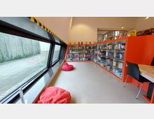 Ein moderner Leseraum mit einer langen Fensterfront auf der linken Seite und Bücherregalen entlang der rechten Wand. Im Raum befinden sich rote Sitzsäcke auf einem hellen Boden und ein Tisch mit einem schwarzen Stuhl im Vordergrund.