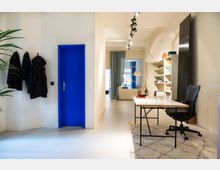 Helles, modernes Büro mit weißen Wänden, einer markanten blauen Tür, einem Schreibtisch mit Bürostuhl auf einem Teppich und Regalen an der rechten Wand. Im Hintergrund befinden sich ein großes Fenster mit halb geöffnetem Vorhang und eine gemütliche Sitzecke.