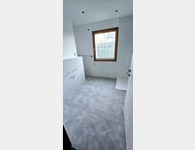 Minimalistisches Badezimmer im Bauzustand mit weißen Wänden und grauen Bodenfliesen. Ein rechteckiges Fenster mit Holzrahmen bietet Ausblick auf eine Hecke, und in der rechten Ecke ist eine flache Duschfläche erkennbar.