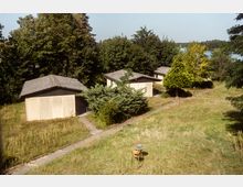 Kleine, flache Gebäude mit Pultdächern stehen inmitten einer Wiese, umgeben von Bäumen und Sträuchern, in einer ländlichen Umgebung. Im Vordergrund befindet sich ein einzelner Stuhl mit einem kleinen Tisch, im Hintergrund ist ein Gewässer zu erkennen.