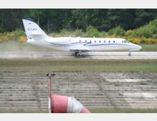 Ein kleines Privatflugzeug rollt über eine betonierte Landebahn mit angrenzendem Grasstreifen, umgeben von Bäumen im Hintergrund. Im Vordergrund ist ein rot-weiß gestreifter Windsack sichtbar.